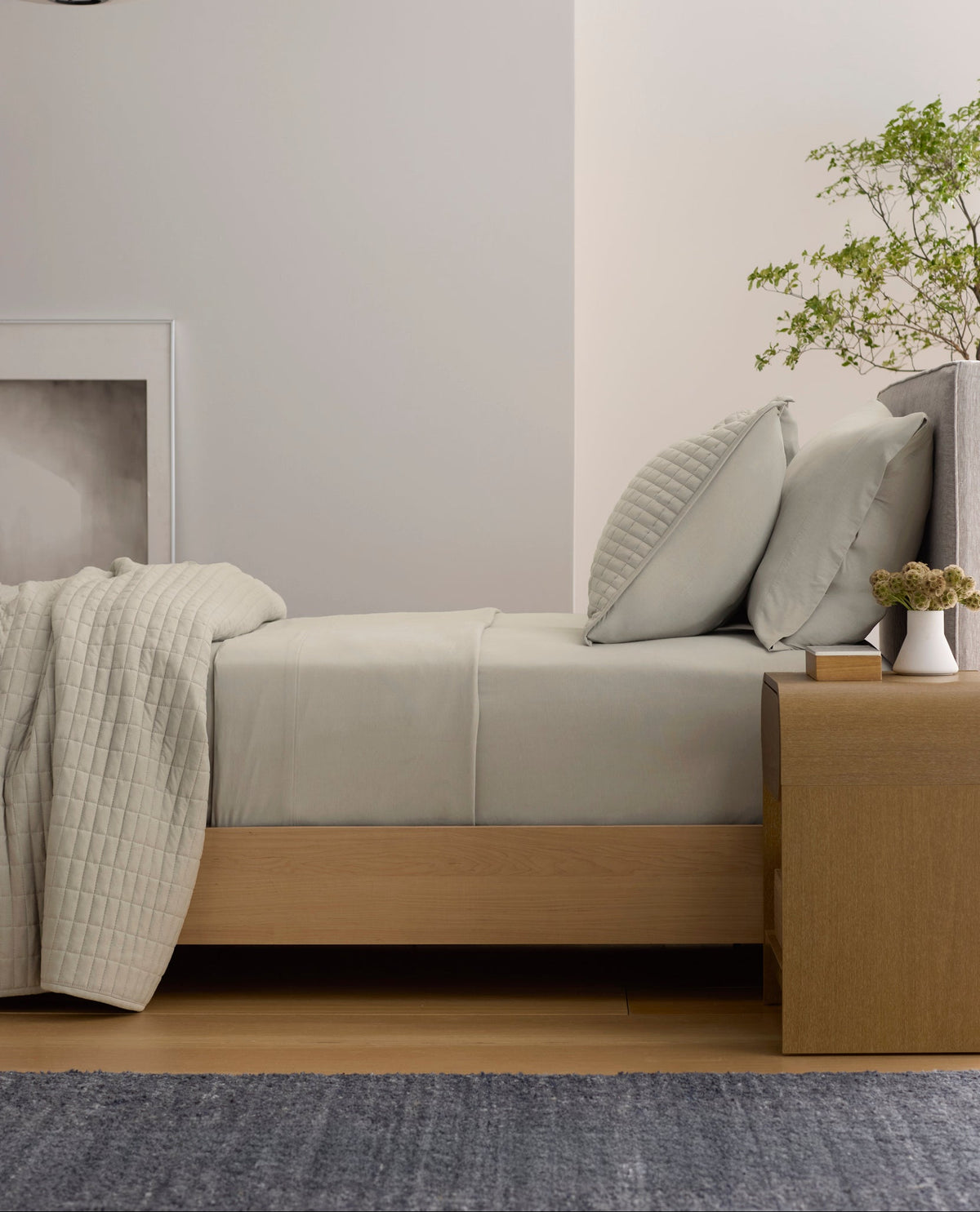 A minimal bedroom features a neatly made bed with Cozy Earth's Bamboo Jersey Sheet Set in pale pink, soft light filtered through sheer curtains, a wooden side table, framed abstract art, and two vases on a wall shelf.|Color:Petal
