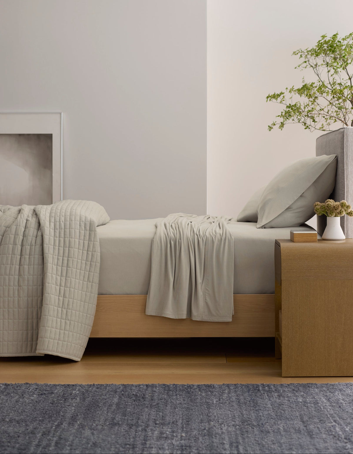 A minimal bedroom featuring Cozy Earth's Bamboo Jersey Sheet Set, a wooden bed, matching quilt, nightstand with plants, modern white pendant lamp, framed artwork on a light wall, and a blue rug on the floor.
