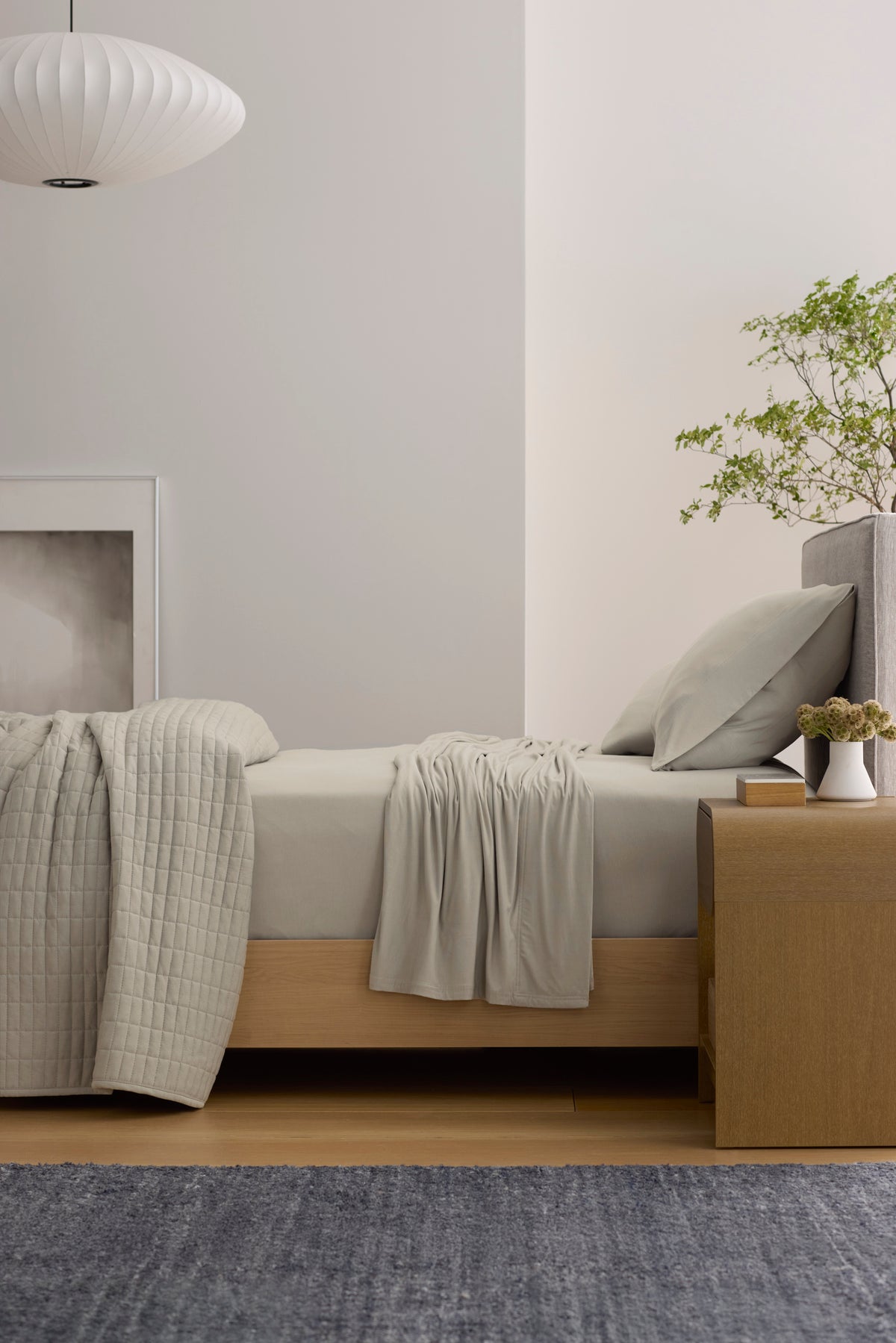 A modern bedroom features a bed with Cozy Earth’s Bamboo Jersey Sheet Set in blue, a light wood nightstand with a cup, books, and lamp, a chair, a plant, sheer curtains, abstract blue wall art, and soft natural light.|Color:Sailor