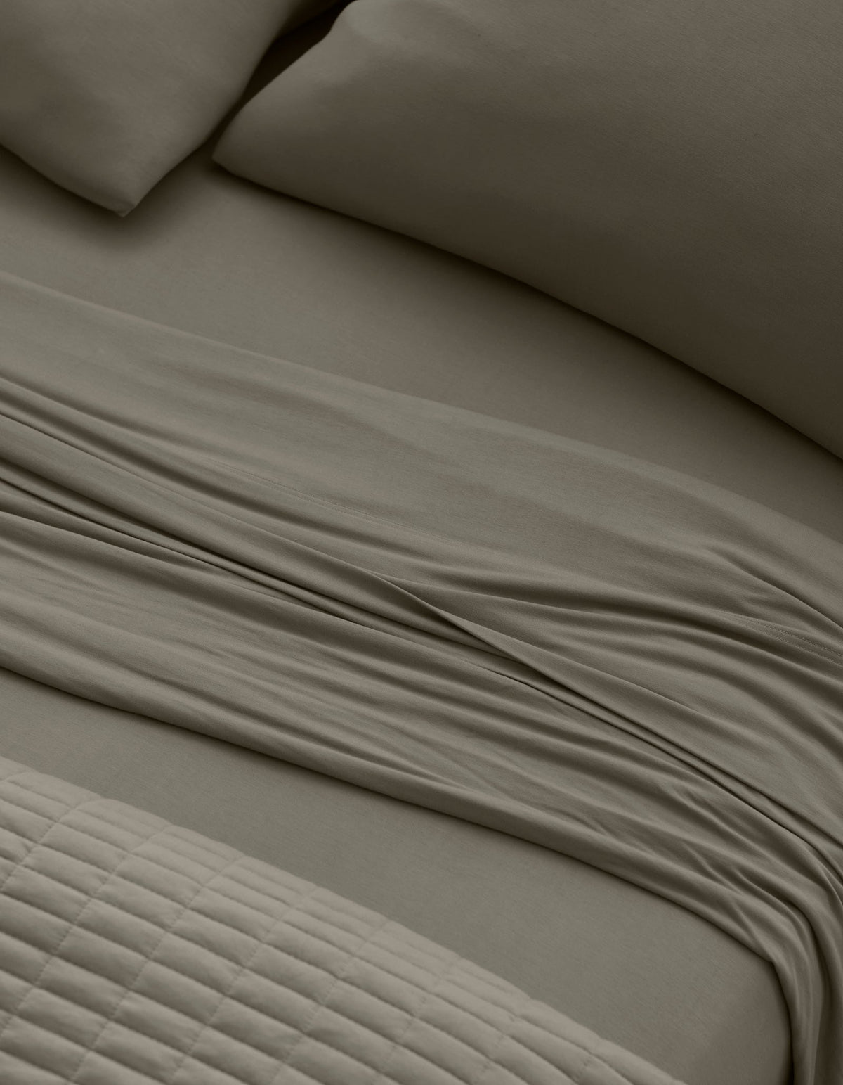 Close-up of a neatly made bed with Cozy Earth Bamboo Jersey Sheet Set in taupe, featuring pillows, a slightly rumpled flat sheet, and a textured quilted blanket at the foot of the bed.