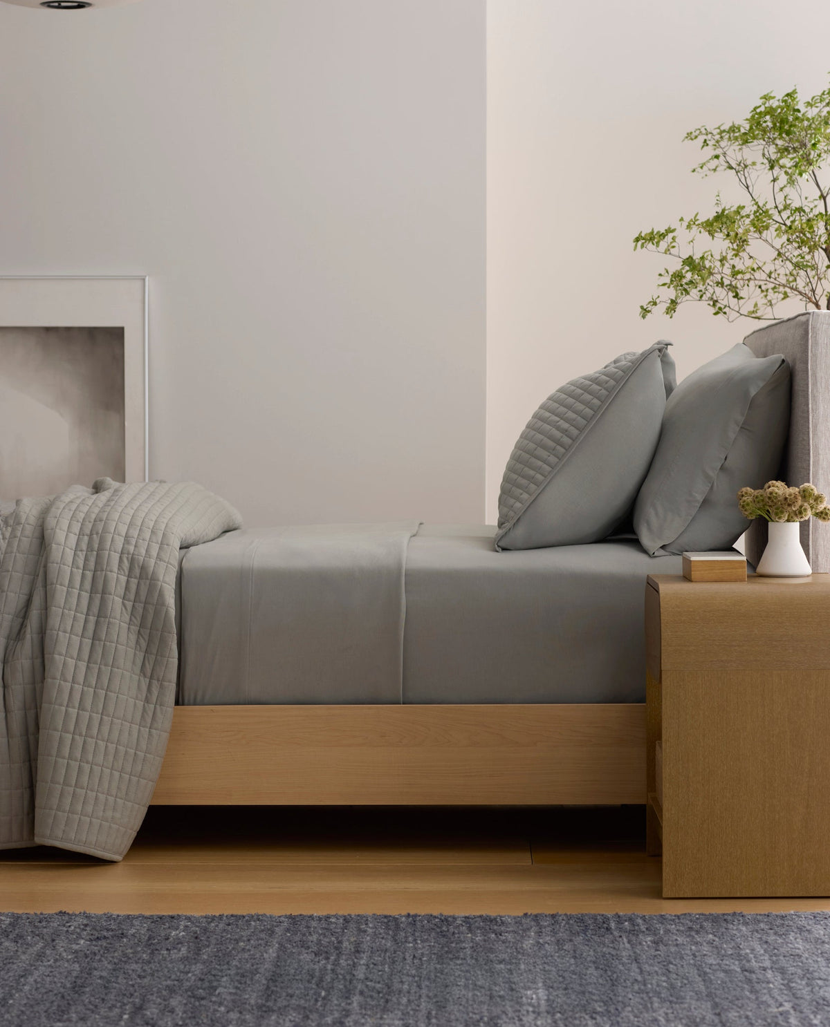 A neatly made bed features the Cozy Earth Bamboo Jersey Flat Sheet in gray, paired with pillows. Next to it, a wooden nightstand holds a potted plant, books, and a vase, while a hanging light and minimalist decor create a calm, modern bedroom.