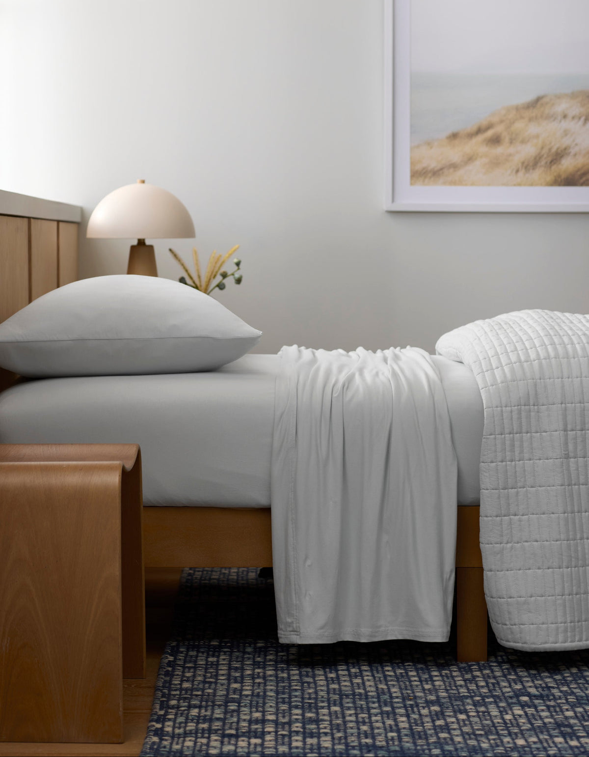 A bed made with Cozy Earth’s Bamboo Jersey Sheet Set rests against a wooden headboard. A wooden bench stands at the foot, while a lamp, small vase, and framed landscape photo accentuate the minimal, serene bedroom.