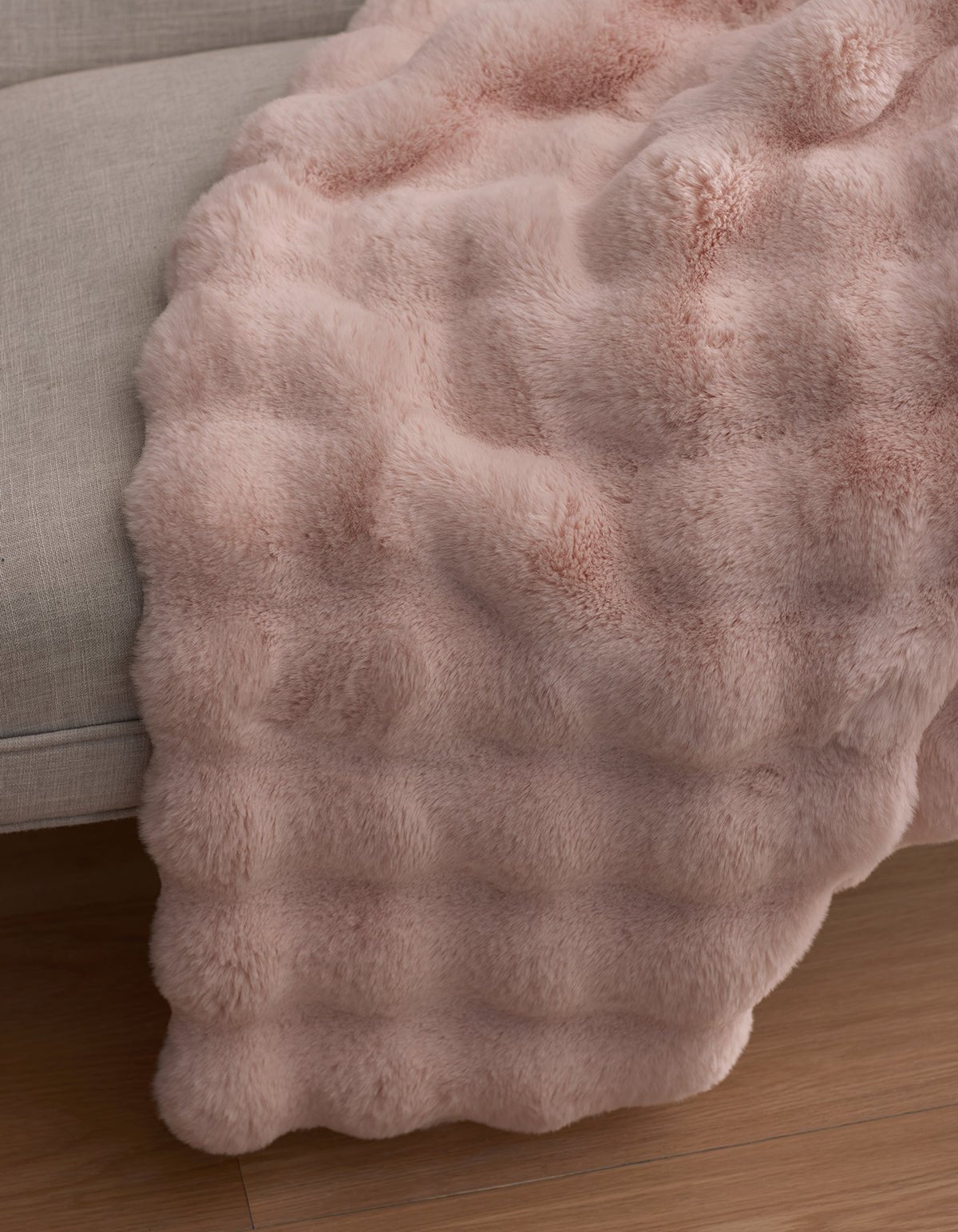 The Cozy Earth Bubble Cuddle Blanket in soft light pink faux fur is draped over the edge of a light gray sofa, cascading gently onto a wooden floor.