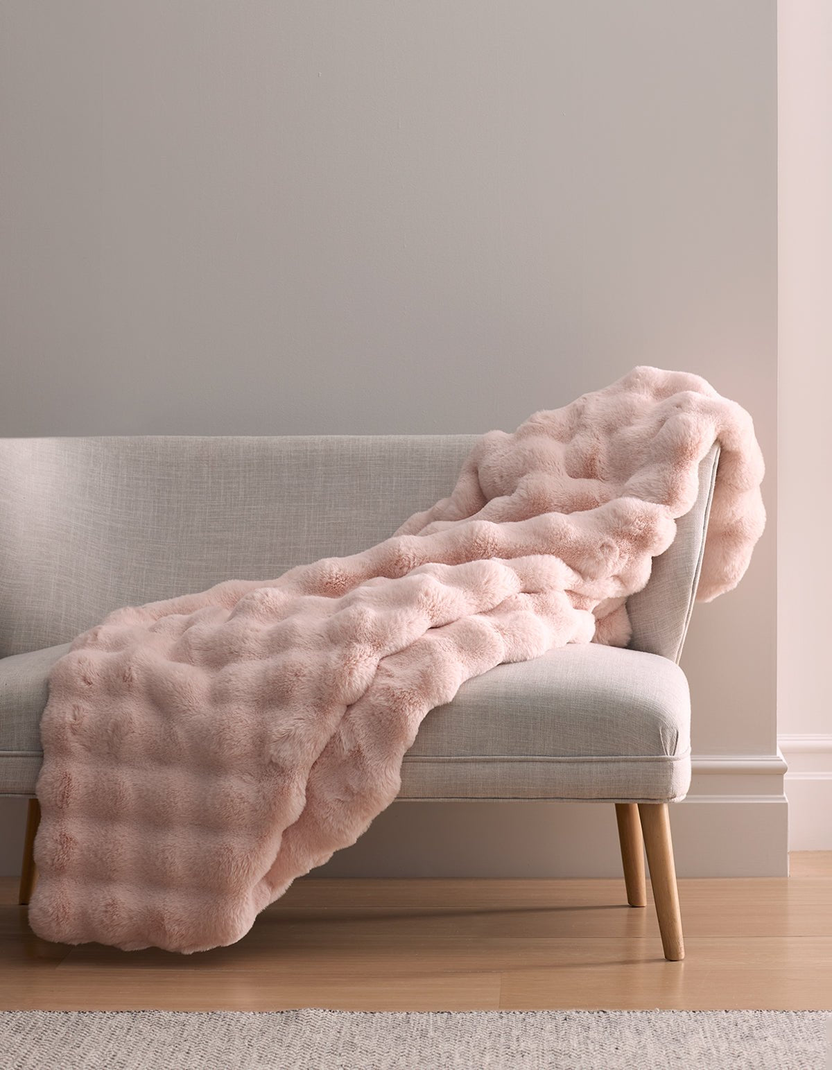 The Bubble Cuddle Blanket by Cozy Earth, in light pink faux fur, is draped over a light gray upholstered bench with wooden legs, against a neutral wall and hardwood floor.|Color:Blossom