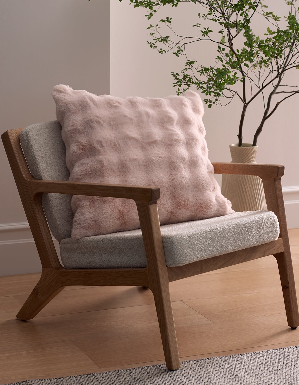 A modern wooden armchair with light gray upholstery sits on a wood floor, featuring the Bubble Cuddle Pillow by Cozy Earth. A leafy green plant in a cream vase stands beside the chair against a pale wall.