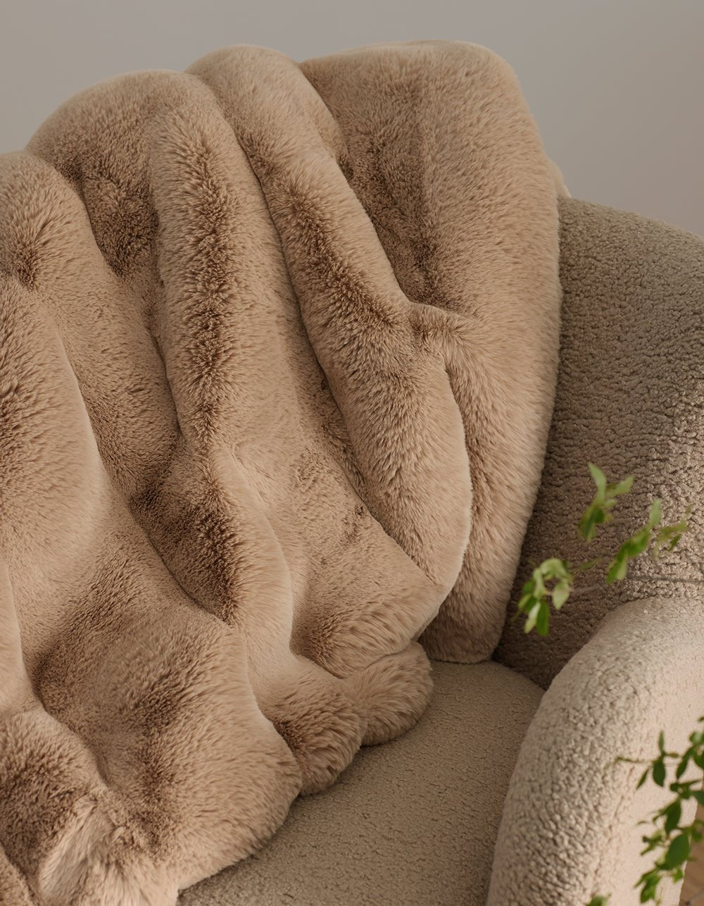 The Channel Cuddle Blanket by Cozy Earth, a plush beige faux fur throw, is draped over a textured light tan armchair. Green leaves peek in from the lower right, adding color to the inviting scene.