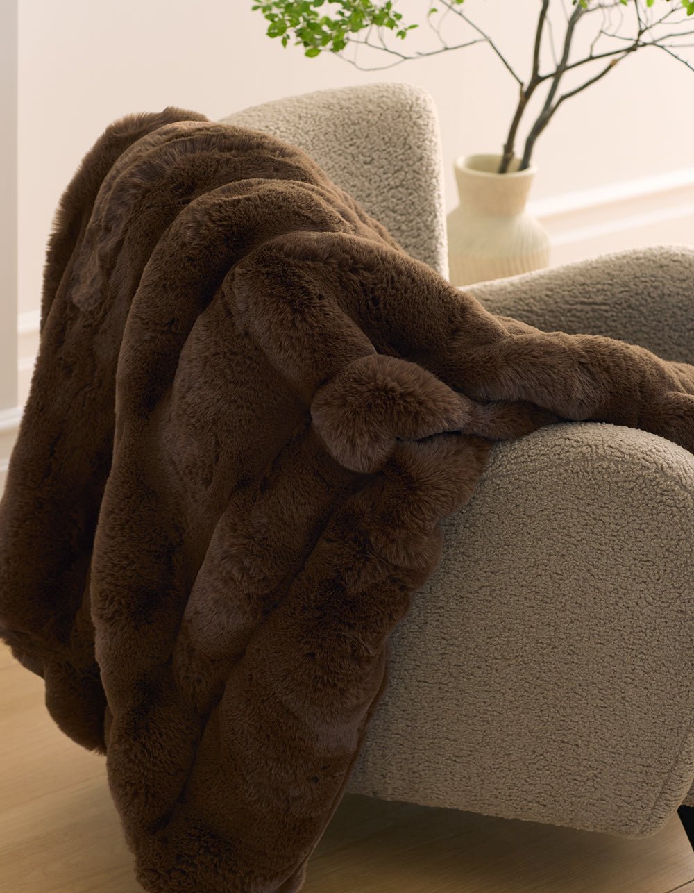 The Cozy Earth Channel Cuddle Blanket, in plush dark brown faux fur, is draped over a textured light beige armchair, with a cream vase of green branches in the background.