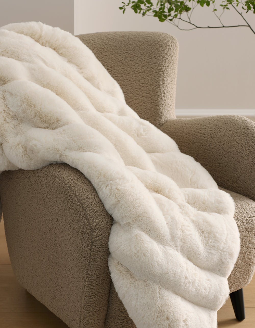 The Cozy Earth Channel Cuddle Blanket, a plush cream-colored faux fur throw, is draped over a textured beige armchair in a cozy modern room with a plant in the background.