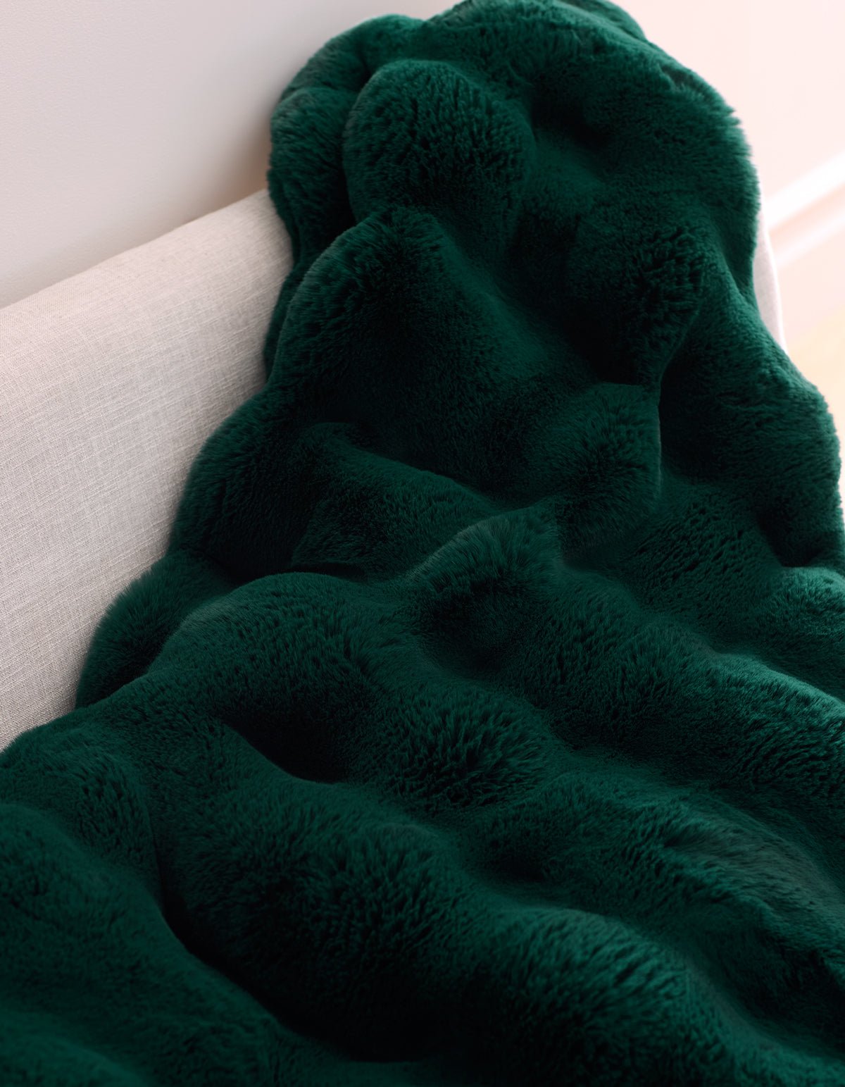 The Double Bubble Cuddle Blanket by Cozy Earth, in dark green plush faux fur with a wavy texture, is draped over the back of a light-colored sofa.