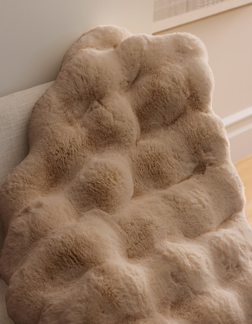 A close-up of the Double Bubble Cuddle Blanket by Cozy Earth, featuring soft ivory beige textured faux fur, draped over a light couch and illuminated by natural light for a cozy look.