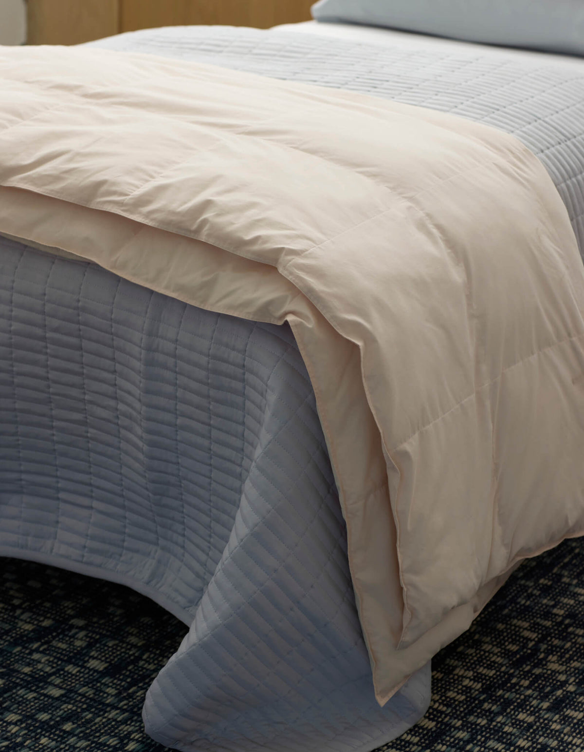 A close-up of a neatly made bed featuring the Cozy Earth Down Alternative Throw, a light blue quilted bedspread, and a beige comforter folded at the foot, all set on a dark patterned carpet.|Color:Coconut