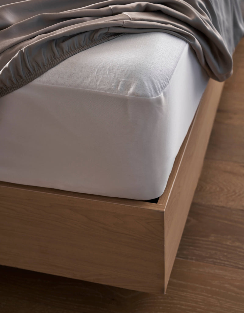 A close-up of a bed with a light wood frame features the Cozy Earth Cooling Mattress Protector beneath a white fitted sheet and gray blanket, all set atop a wooden floor.