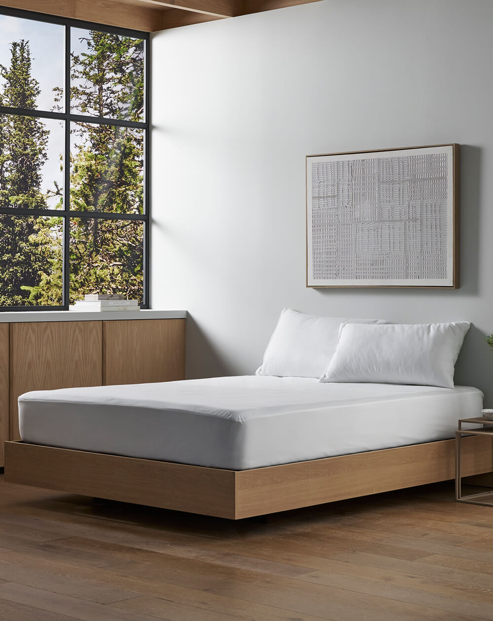 A modern bedroom featuring Cozy Earth’s Cooling Mattress Protector on a wooden platform bed with white bedding, a large window with green views, wood floors, minimalist wall art, and built-in wood cabinetry.