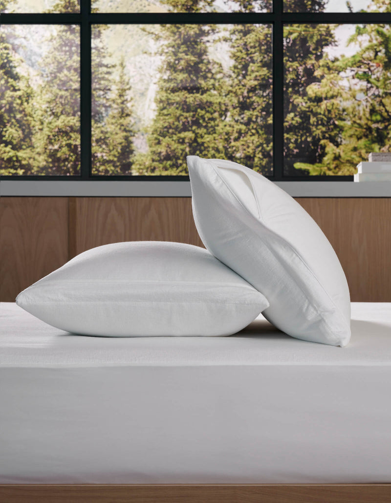 Two Cozy Earth Cooling Pillow Protectors are stacked on a neatly made bed by a large window with views of green trees, complementing the room's modern, minimalistic design.