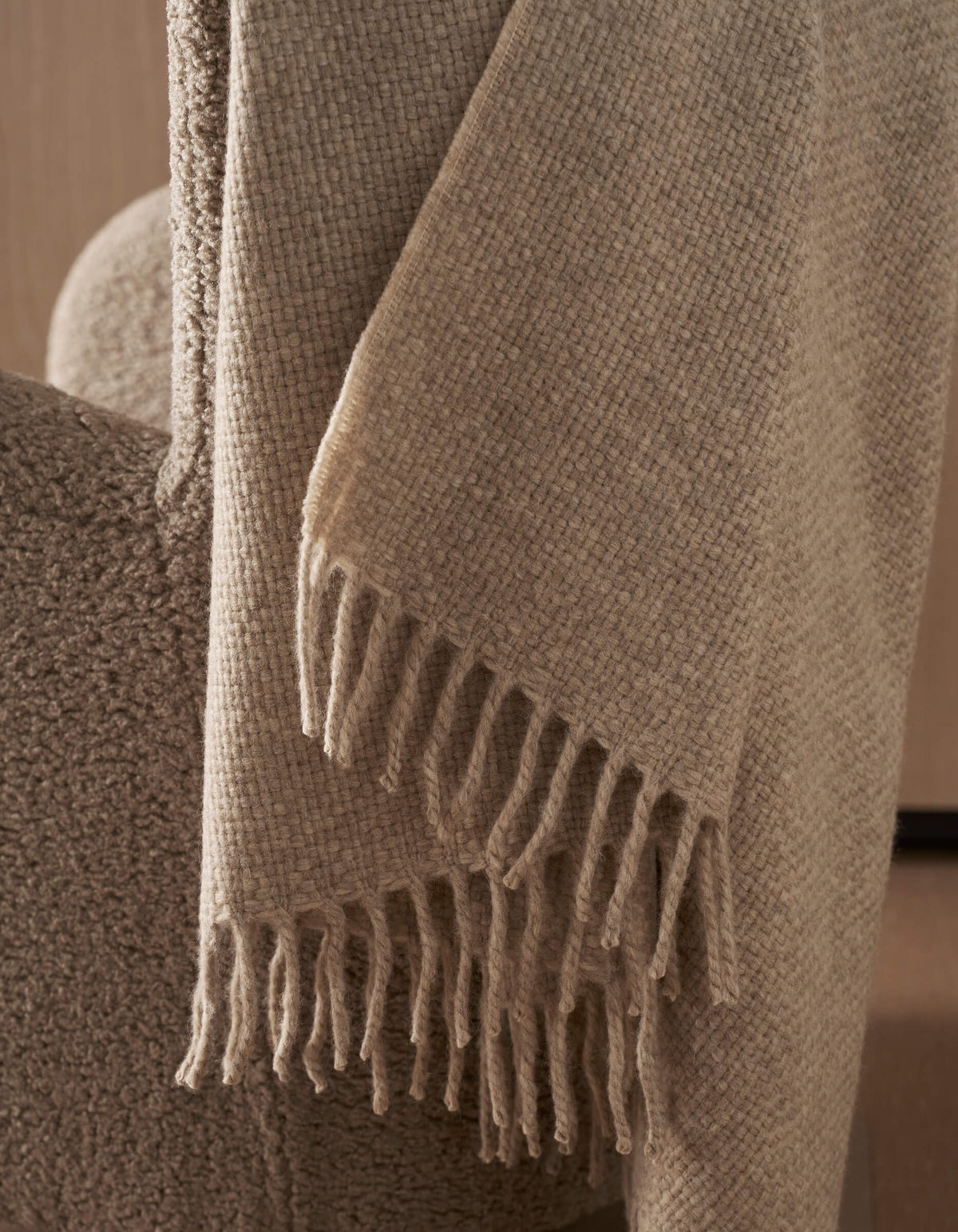 The Cozy Earth Lucia Throw, featuring a beige textured design with fringe, drapes over the arm of a soft upholstered chair in a neutral-toned room.