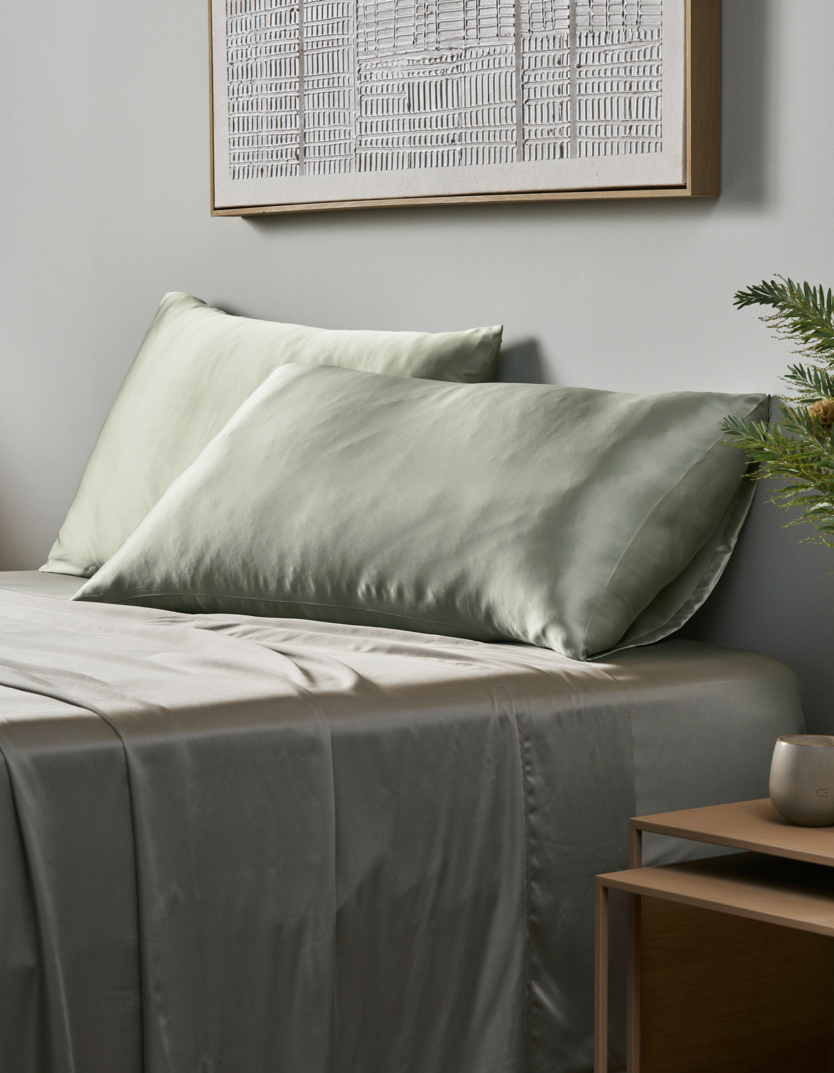 A neatly made bed features Cozy Earth Silk Pillowcases in light sage green, complemented by a wooden nightstand with a plant and cup. A framed abstract artwork decorates the light gray wall above.