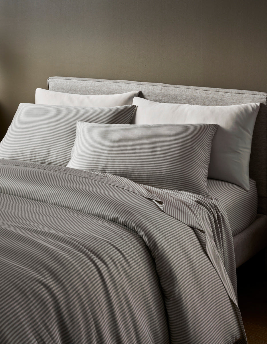 A neatly made bed with a light gray upholstered headboard, white pillows, and the Cozy Earth Bamboo Duvet Cover in gray stripes sits in a softly lit room with neutral walls.