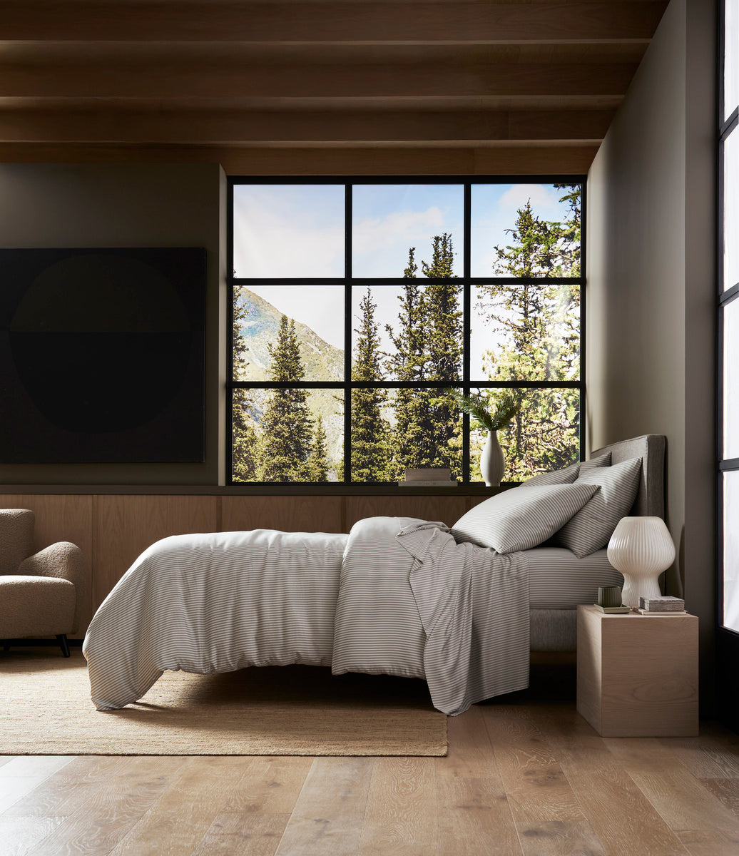 A modern bedroom features a neatly made bed with Cozy Earth's Bamboo Duvet Cover, light bedding, a nightstand and lamp, wood floors, and large windows overlooking pine trees and mountains under a blue sky filled with natural light.
