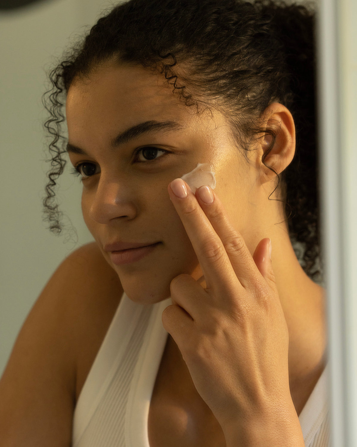 A person with curly hair is applying Cozy Earth's Water Cream Moisturizer with their fingers, concentrating on their left cheek. Their expression is calm and focused. They are wearing a sleeveless white top, and the background is softly blurred. 