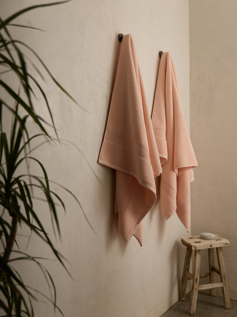 Two Cozy Earth Luxe Bath Sheets in soft pink hang neatly on hooks against a neutral wall. Below, a rustic wooden stool holds a neatly folded cloth. A leafy plant partially visible on the left adds a touch of greenery.