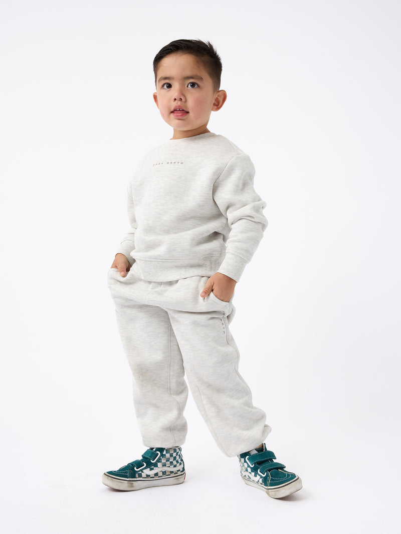 A young child stands confidently against a plain background, dressed in the Kid's CityScape Crewneck by Cozy Earth and sporting green sneakers. With hands tucked into pockets, the child strikes a relaxed pose. The outfit looks comfortable and casual.