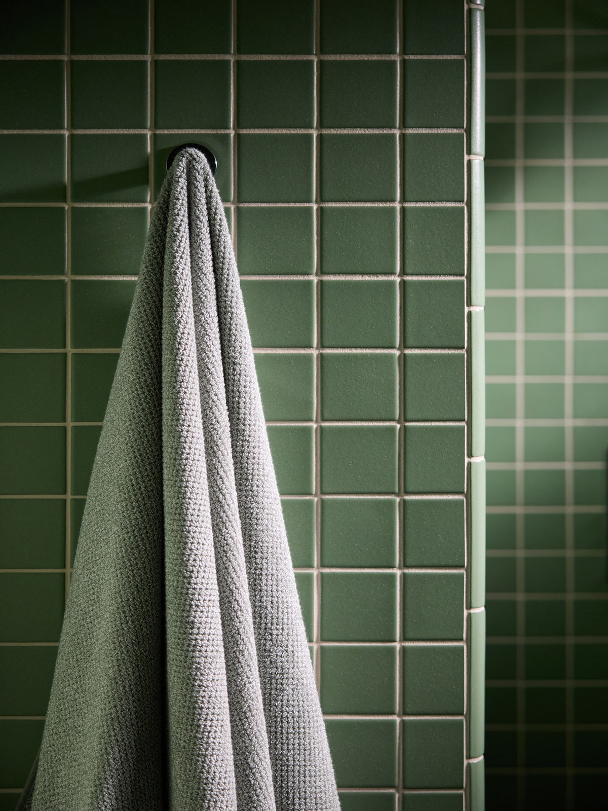 A Nantucket Bath Towel in Heathered Sage is shown hanging in a green tiled bathroom. 