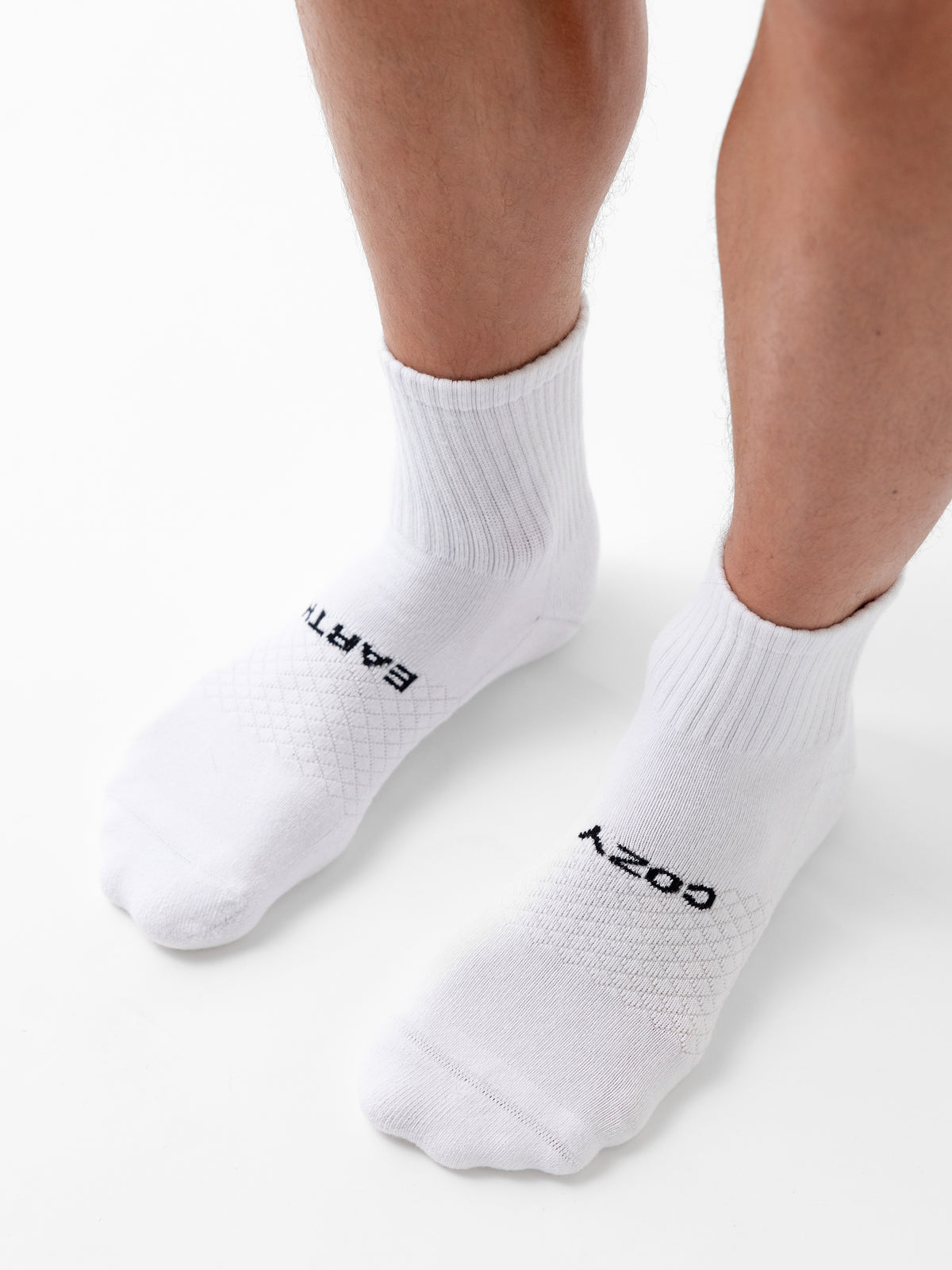 Someone stands on a white surface wearing Cozy Earth’s Essential Quarter Socks featuring ribbed cuffs and black "EARTH" text on the left sock and "COZY" on the right. 