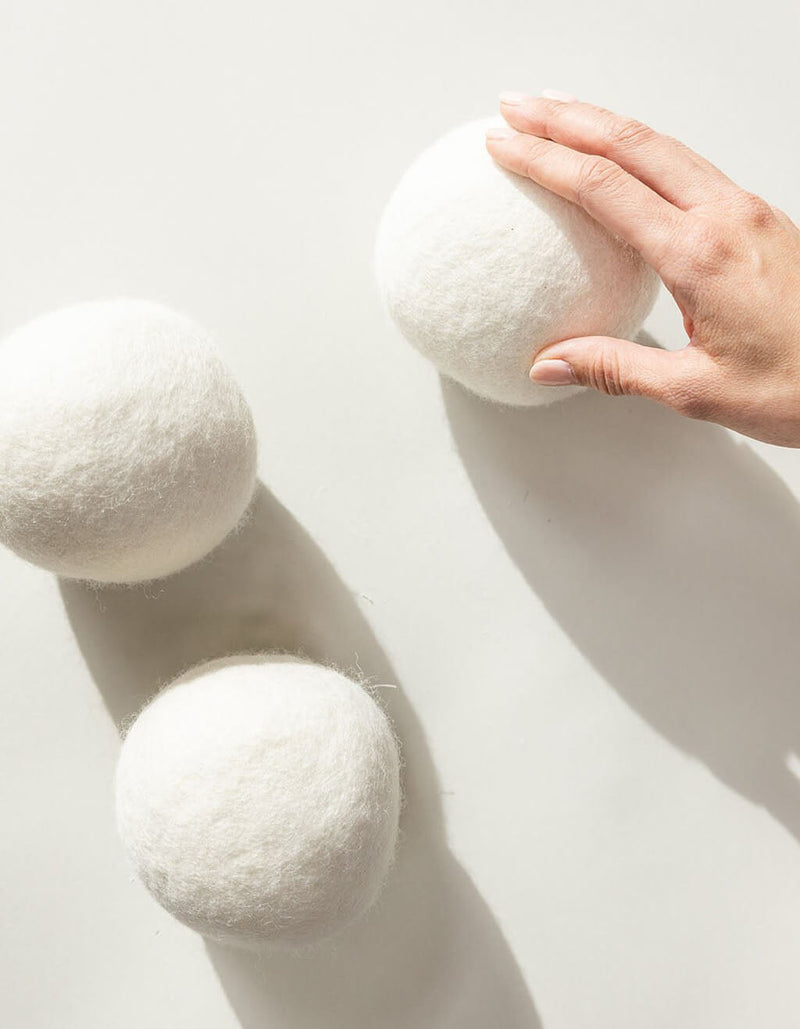 A hand reaches for one of three large Cozy Earth Wool Dryer Balls, displayed on a smooth, light background with soft shadows.