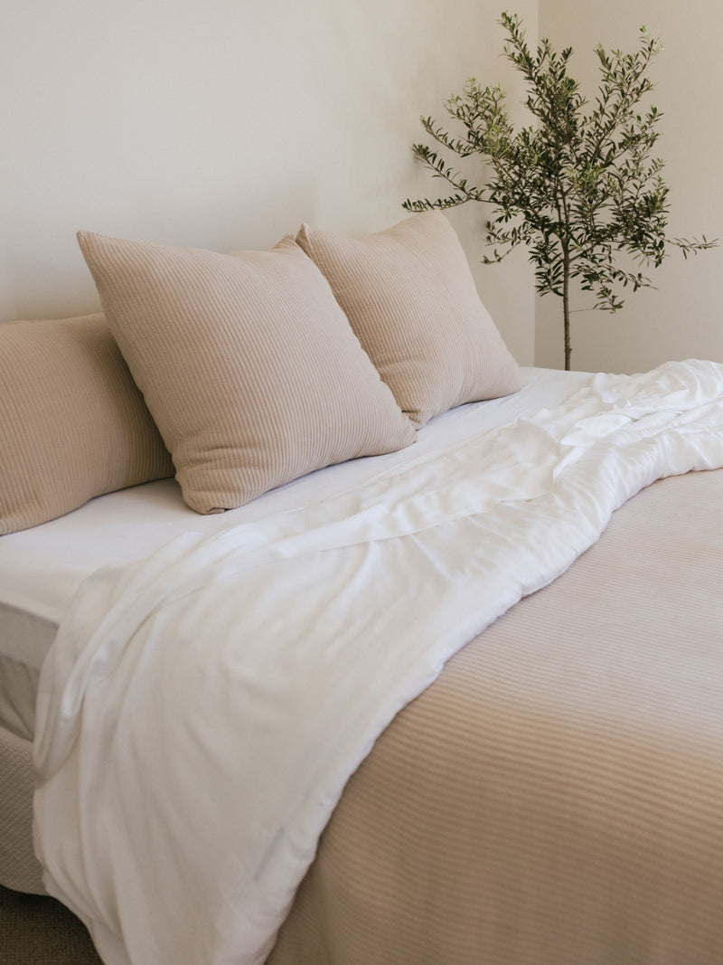 A neatly made bed featuring a light beige Premium Down Alternative Insert by Cozy Earth and three puffy beige pillows, against a light-colored wall. A white sheet is partially draped over the bed. A small green plant in the corner adds a touch of nature to the serene bedroom setting.