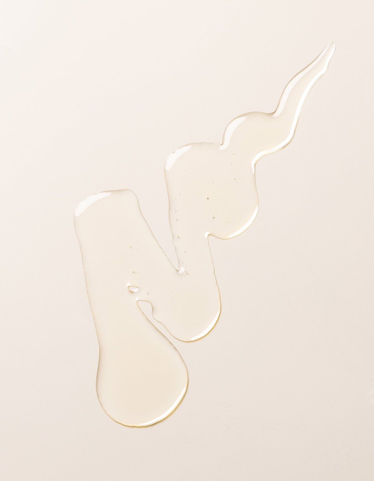 A swirl of transparent, light-colored Cozy Earth's Revitalizing Pair liquid on a plain, pale beige surface. The liquid forms irregular shapes with droplets at the ends, creating a visually interesting pattern.