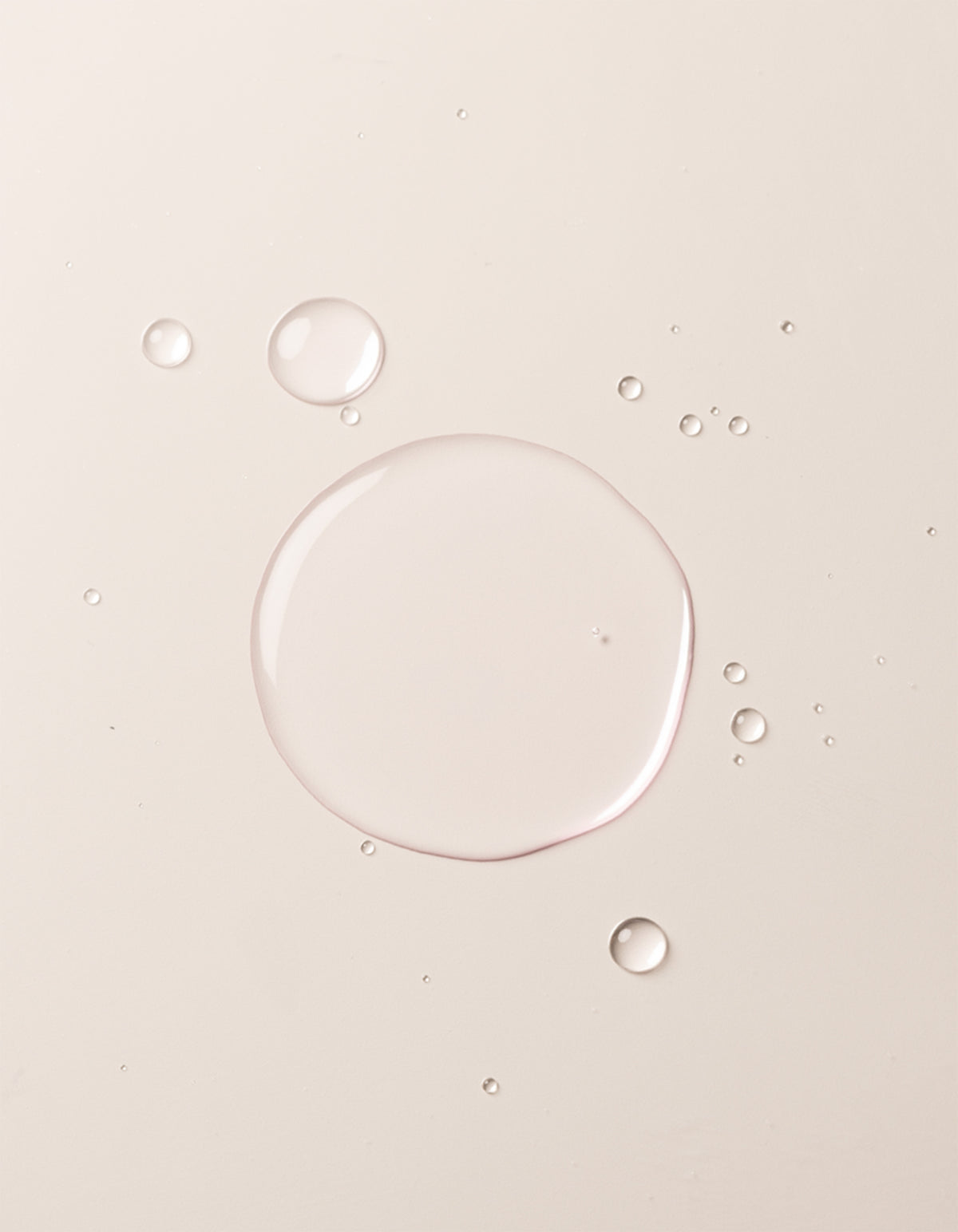 Clear liquid droplets of Balancing Face Mist by Cozy Earth are scattered on a smooth, light beige surface. The largest droplet is centered, surrounded by smaller drops, giving the image a minimal, clean, and soft appearance. 