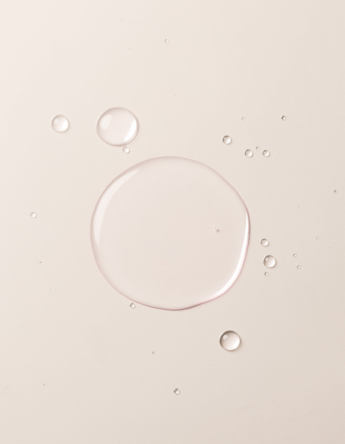 A close-up image showcases translucent liquid droplets of Cozy Earth's Balancing Face Mist on a smooth, off-white surface. The droplets vary in size, with one large central droplet surrounded by several smaller ones, creating a clean and minimalistic appearance. 