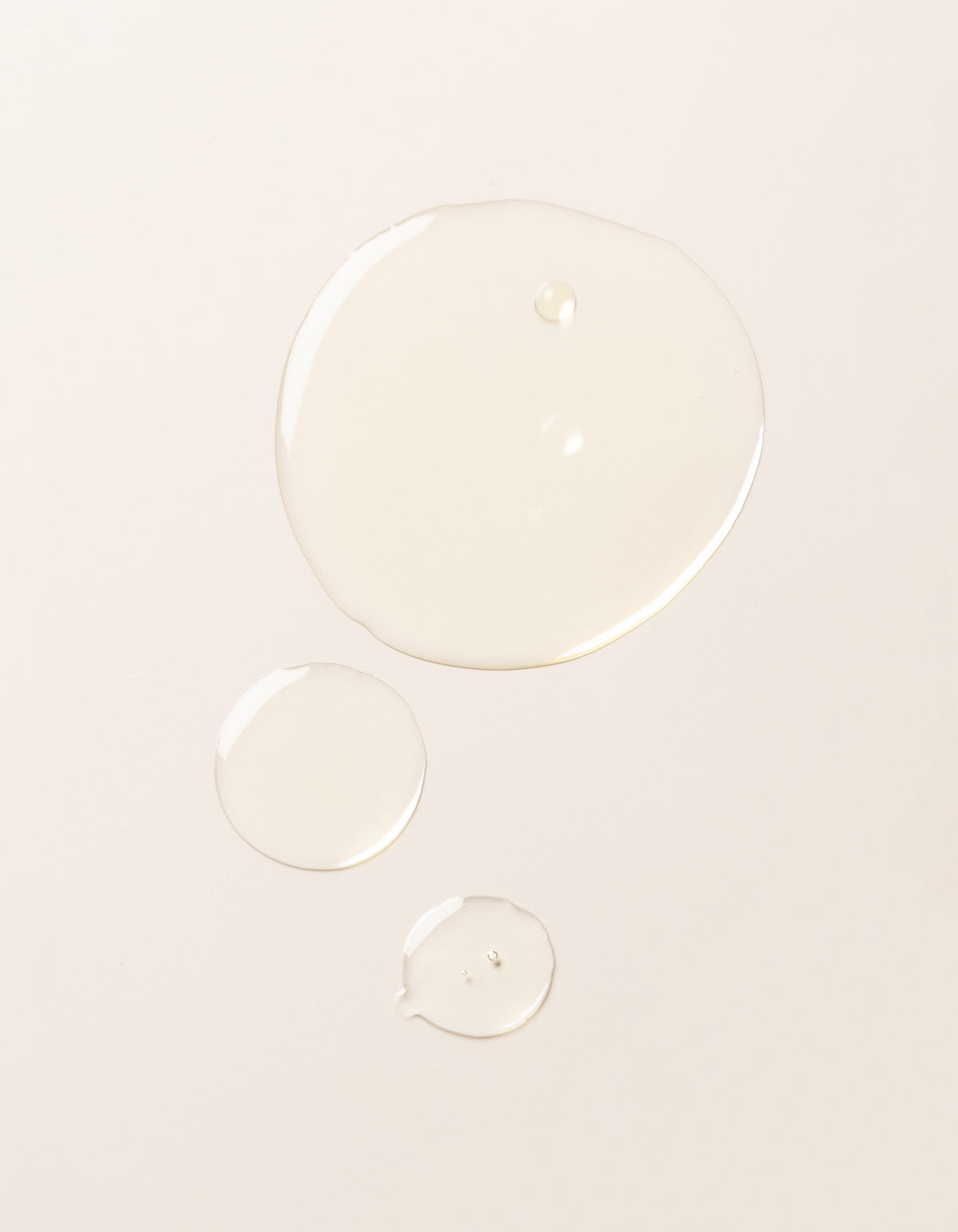 Three droplets of Cozy Earth's Brightening Vitamin C Oil, each of a different size, are spaced apart on a smooth, light-colored surface.