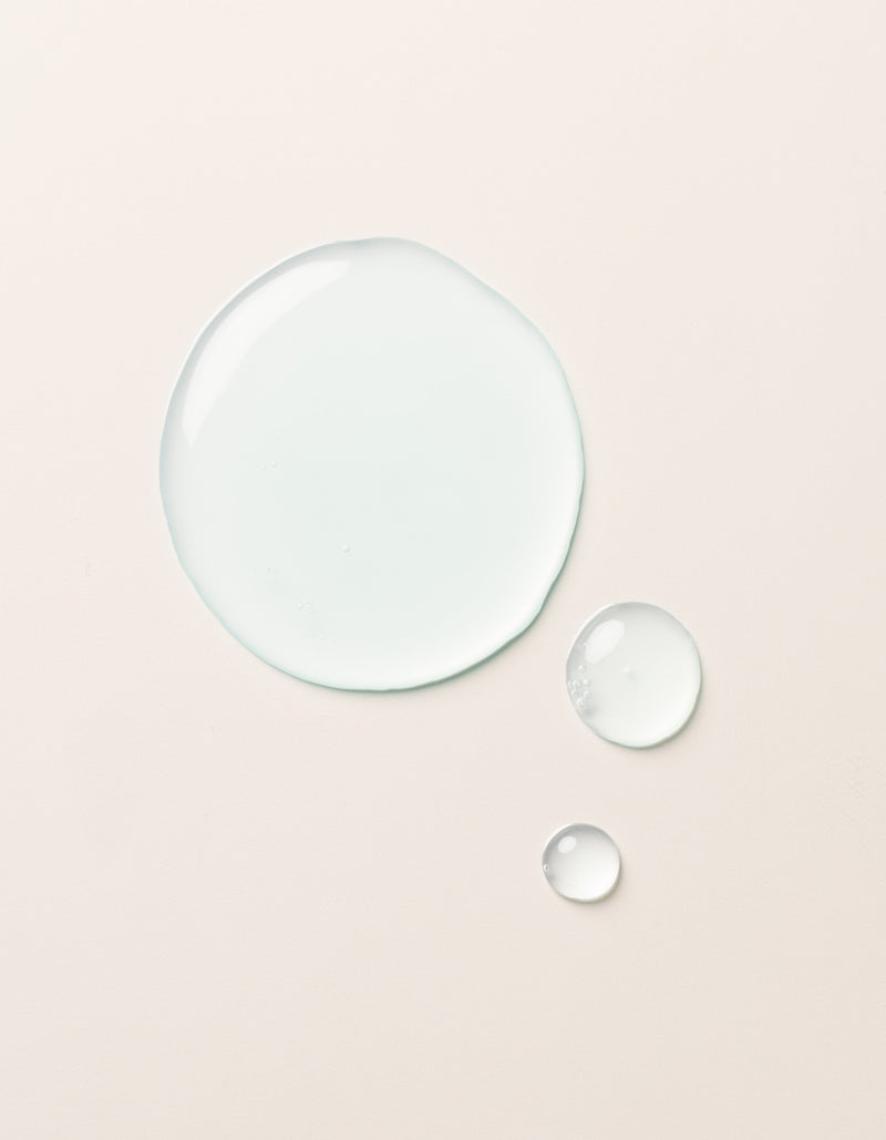 The Hydration Duo by Cozy Earth is shown as a large drop and two smaller drops of clear, light blue liquid on a smooth, light beige surface. The drops vary in size but all maintain a rounded shape, appearing glossy and slightly reflective.