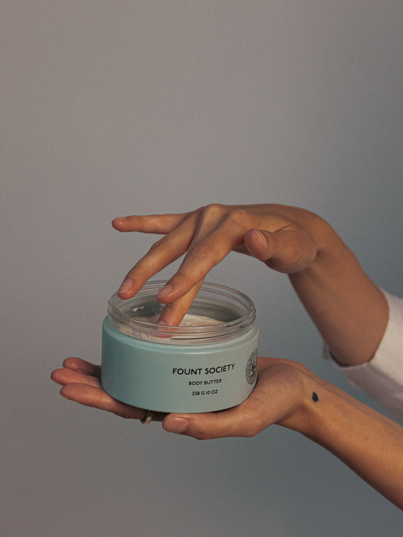 A person dips their fingers into an open jar of "Cozy Earth" body butter, held in their other hand. The jar is teal with black text, and the person's hands are positioned against a neutral background. A small tattoo is visible on their wrist.