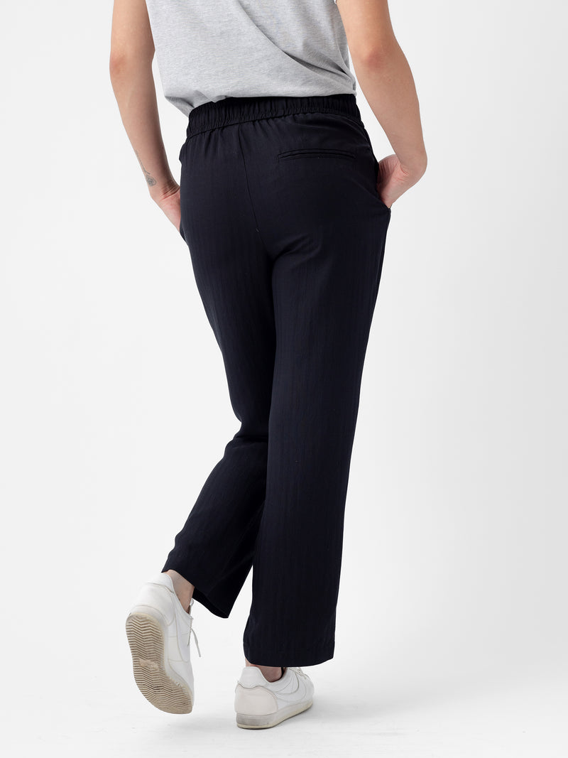 A person is shown from the back, standing against a white background. They are wearing a grey shirt and Cozy Earth's Women's Sunset Cropped Pant, which features an elastic waistband, along with white sneakers. The person's right hand is in their pocket, and they are slightly leaning back.