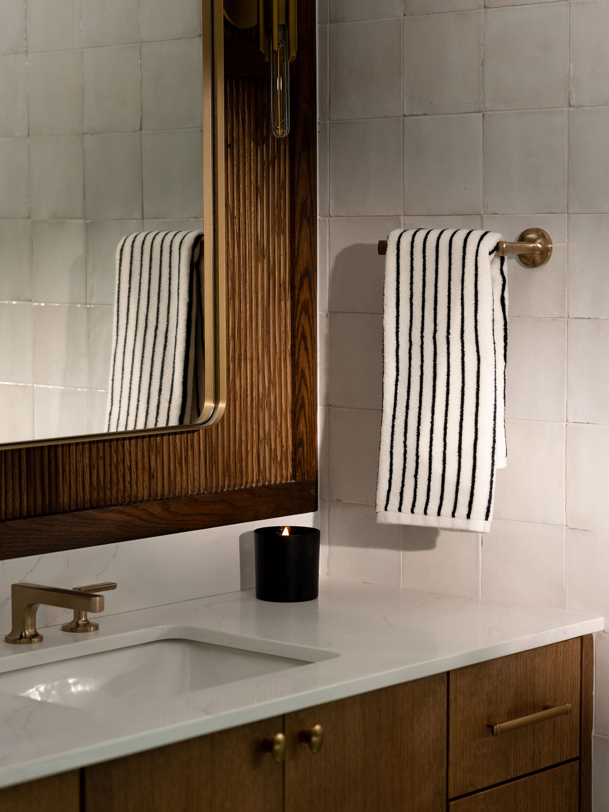 A bathroom scene featuring a white countertop with a sink, brass faucet, and wooden cabinets. A black candle and Luxe Hand Towels by Cozy Earth, hanging on a brass towel bar, are also present. The background consists of white tiled walls and a large wooden-framed mirror. 