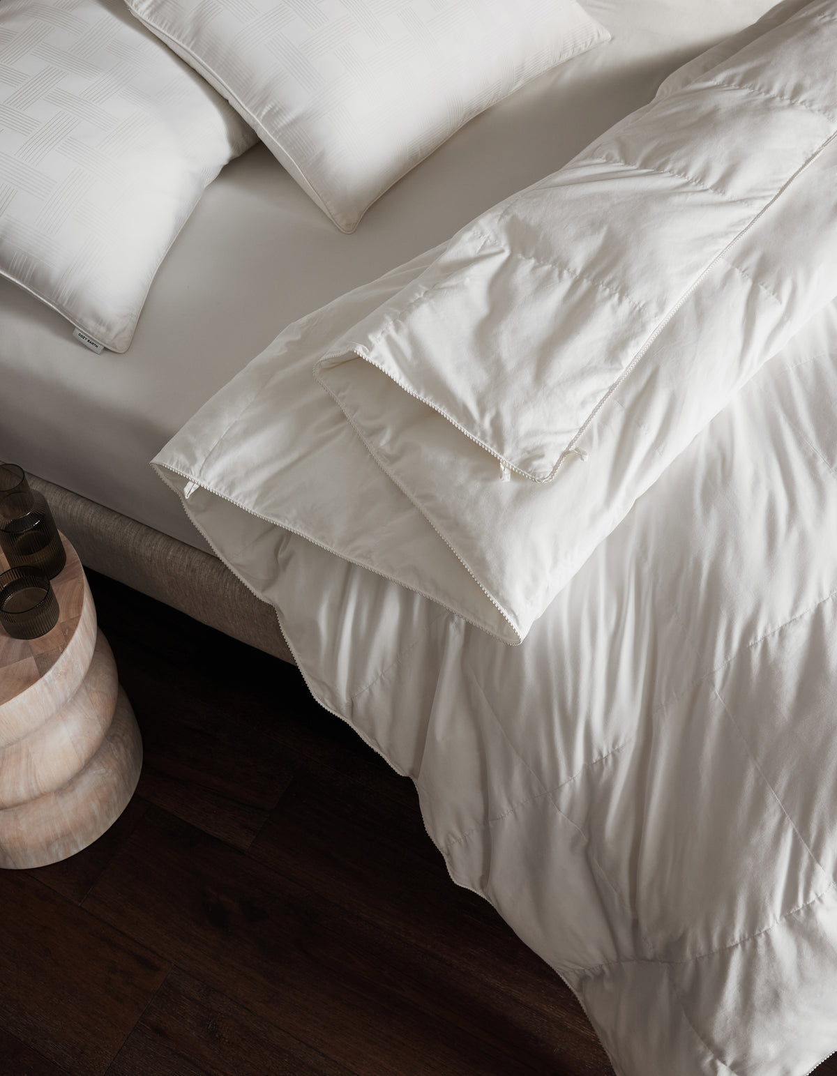 A neatly made bed features Cozy Earth's Down Alternative Comforter and white pillows beside a small wooden nightstand on a dark wood floor.