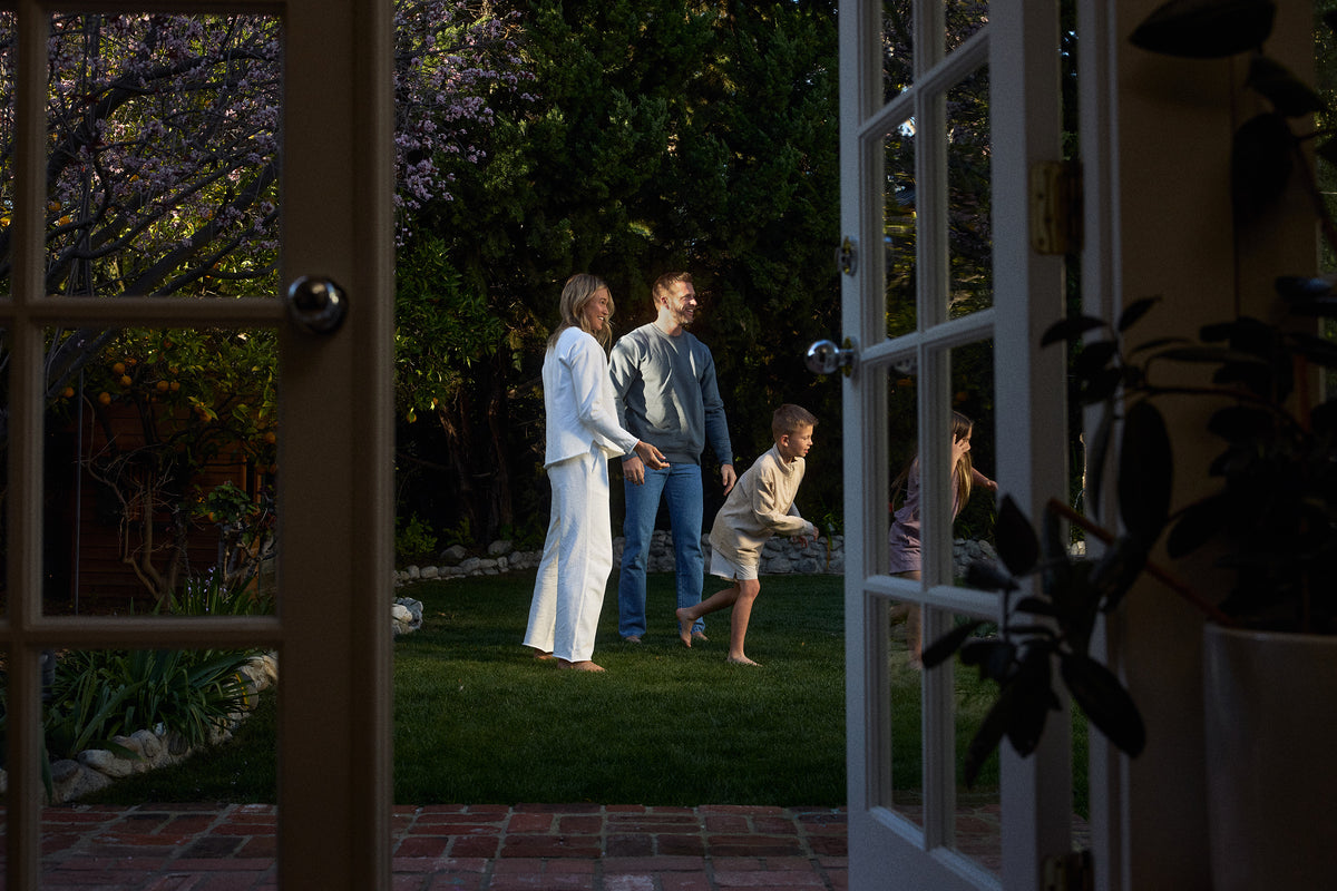 Family standing in a garden with a child playing, viewed through an open door.