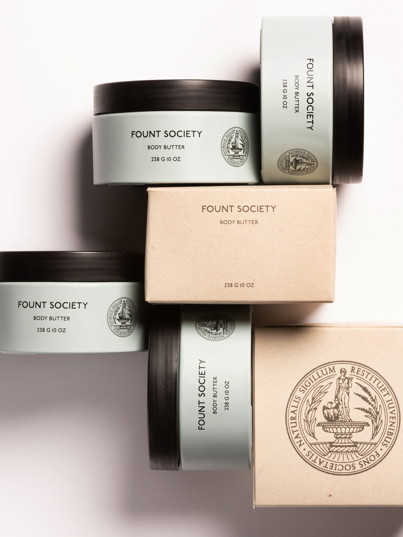 The image features an arrangement of body butter containers from the brand "Cozy Earth." The containers, labeled "Body Butter" with a quantity of 236 g (8 oz), are stacked and include a mix of uncovered and boxed items, showcasing the product branding and logo.
