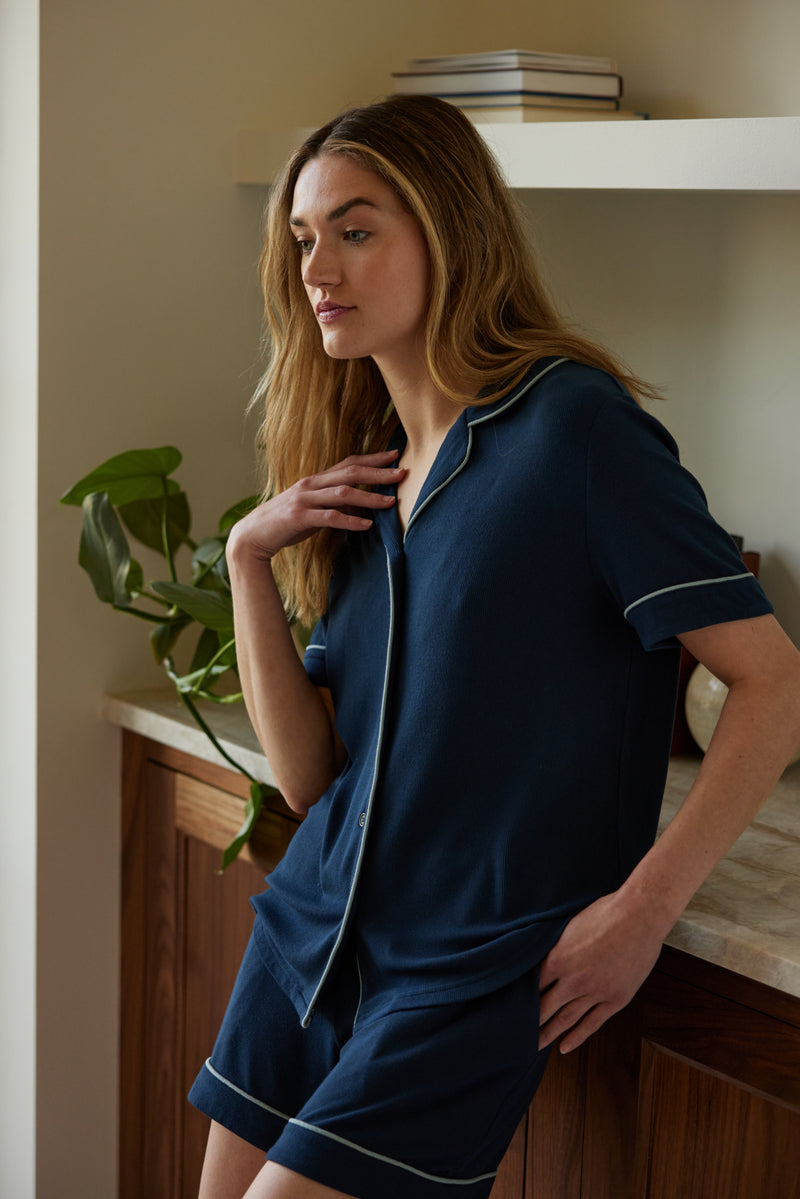 A woman with long hair leans against a wooden counter, wearing the Cozy Earth Women's Bamboo Rib Knit Classic Short Sleeve Pajama Top in navy blue with white piping. She gazes thoughtfully to the side, resting one hand on her chest and the other in her pocket. A potted plant and books are visible in the background.