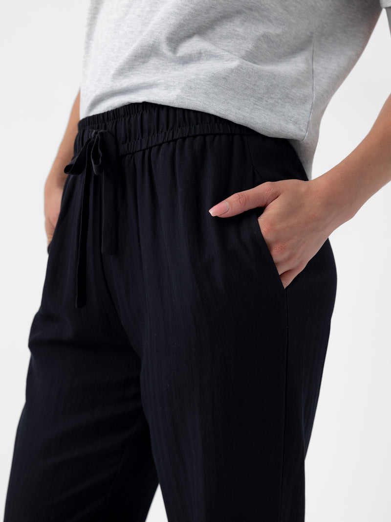 Close-up of a person wearing a casual outfit. They have on a gray t-shirt and Cozy Earth's Women's Sunset Cropped Pant in black, with their right hand inside the pants pocket. The background is plain and white.