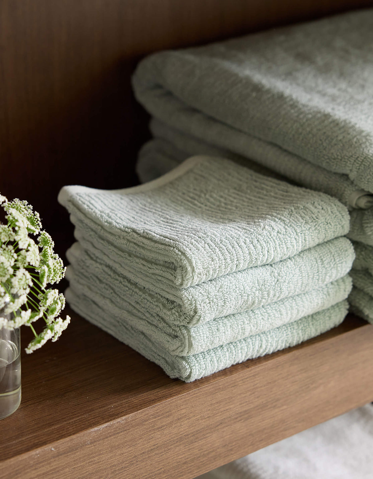 A neatly folded stack of Cozy Earth's Ribbed Terry Washcloths in pale green is placed on a wooden shelf. Beside the washcloths, a small clear glass with a few delicate white flowers in water adds a touch of elegance to the display. 