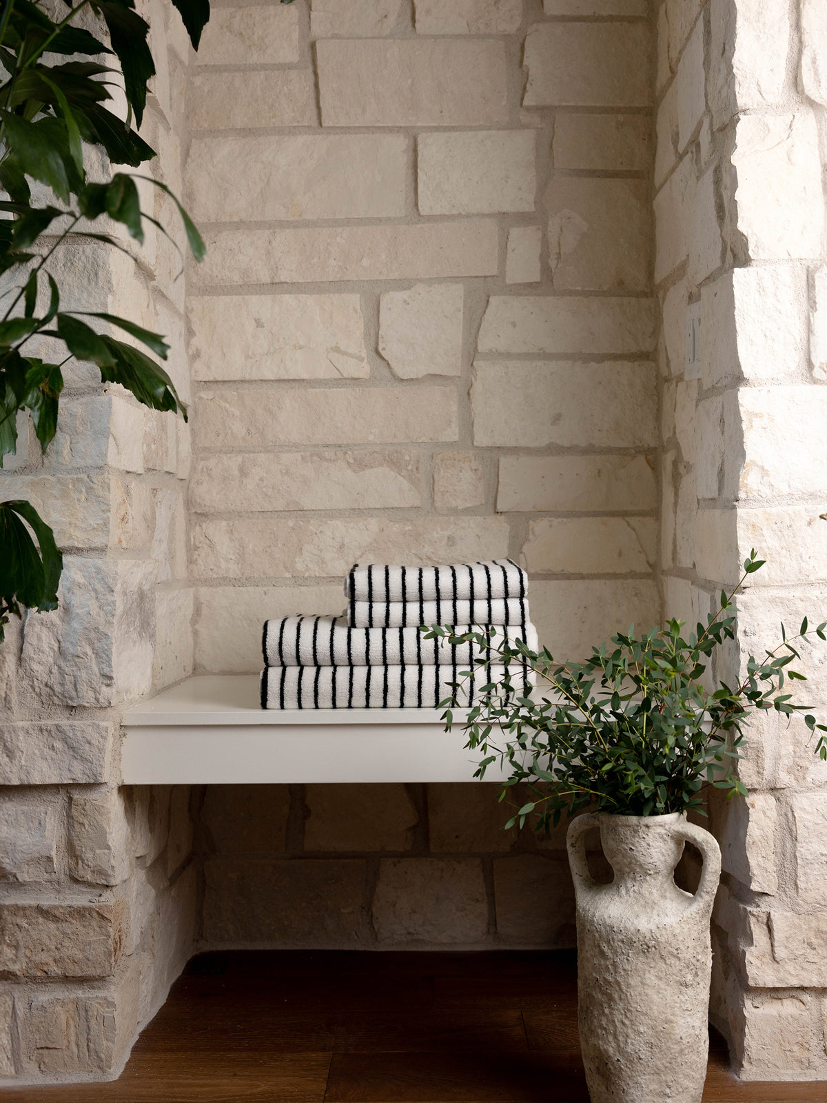 A neatly stacked pile of folded Luxe Hand Towels by Cozy Earth, featuring black and white stripes, sits on a white shelf against a textured stone wall. A rustic vase filled with green leafy branches is placed on the floor next to the shelf. A large plant with dark green leaves is partially visible on the left. 