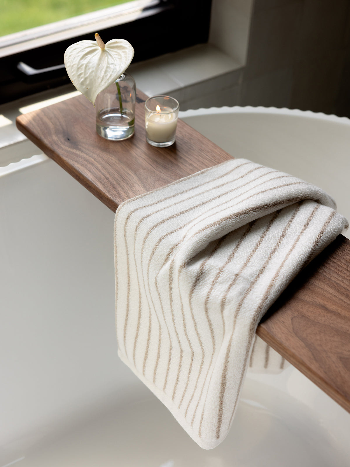 A cozy bathroom setting featuring a wooden bathtub tray with a white anthurium flower in a clear vase, a lit candle, and a folded Luxe Hand Towel by Cozy Earth, striped in white and beige, draped over the tray. The bathtub is white and positioned near a window with greenery outside. 