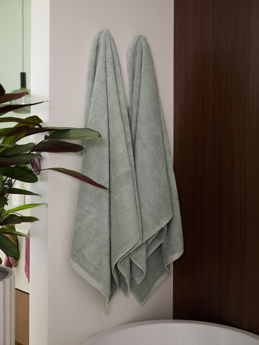 Two Cozy Earth Ribbed Terry Bath Towels in light gray hang side by side on a white wall near a wooden panel, with a green potted plant and part of a bathtub visible in the modern bathroom.