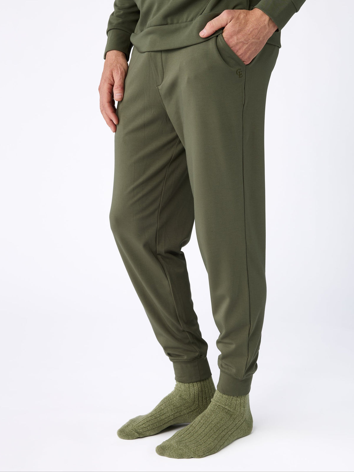 A person dressed completely in Cozy Earth's Men's Ultra-Soft Bamboo Pullover Crew in green stands against a white background. They have their hands in the pockets of their olive green pants and are wearing matching green socks. 