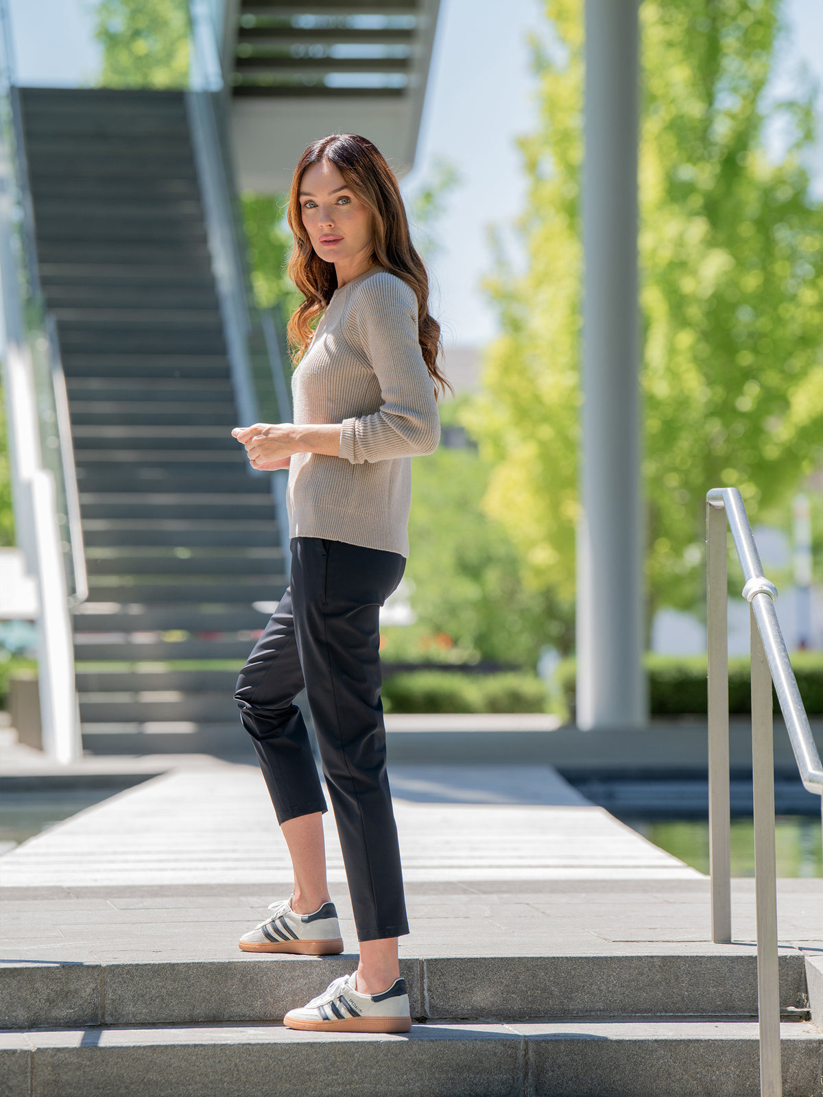 A woman with long brown hair stands on an outdoor staircase, wearing a beige Women's Classic Crewneck by Cozy Earth, black pants, and gray Adidas sneakers. She looks over her shoulder with a serious expression. The background features a modern structure and green trees. 