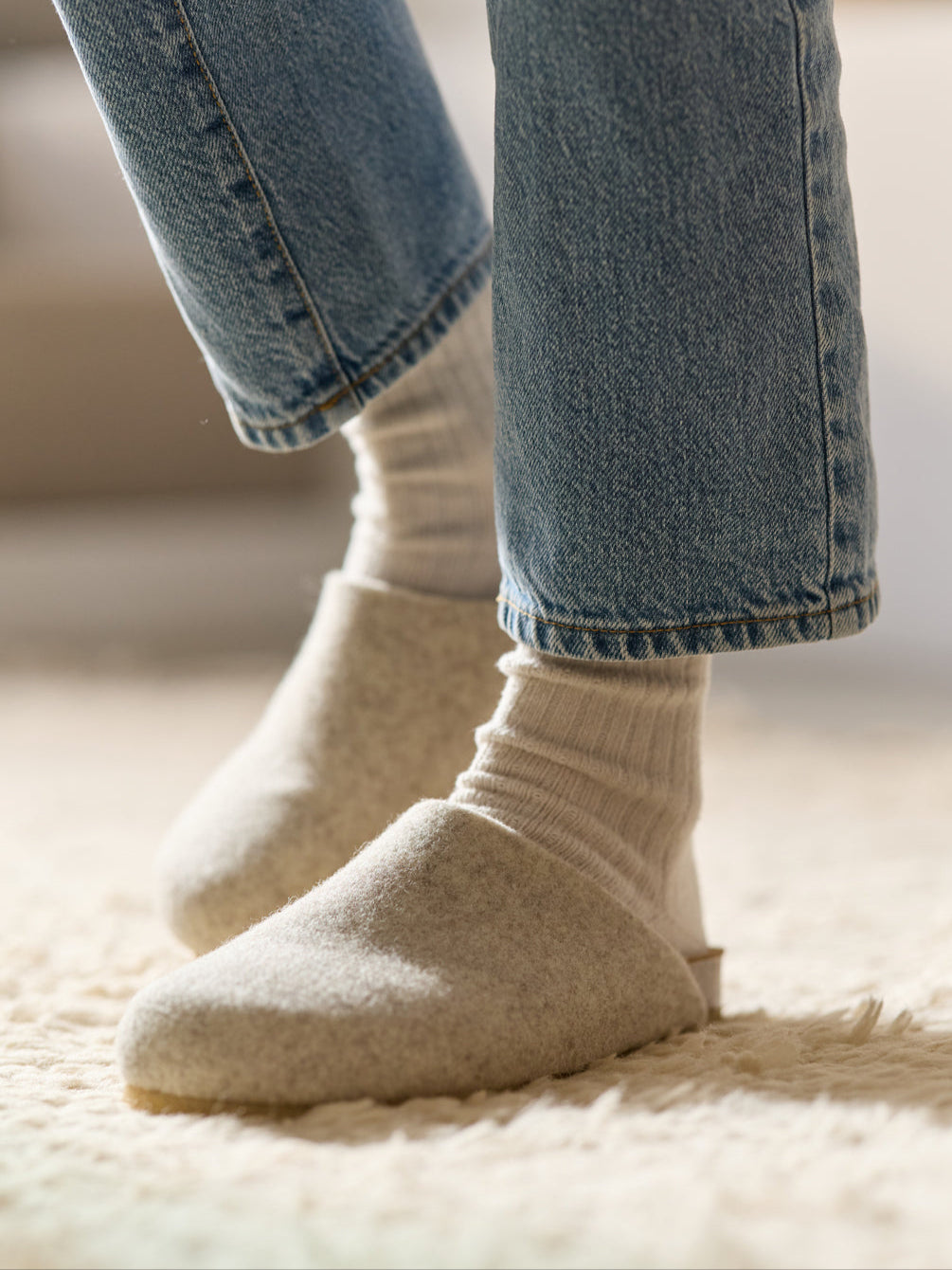 Close-up of a person wearing light-colored felt slippers and white socks, standing on a textured, light-colored rug. They are also wearing blue jeans that gently rest on the slippers. The scene conveys warmth and comfort.
