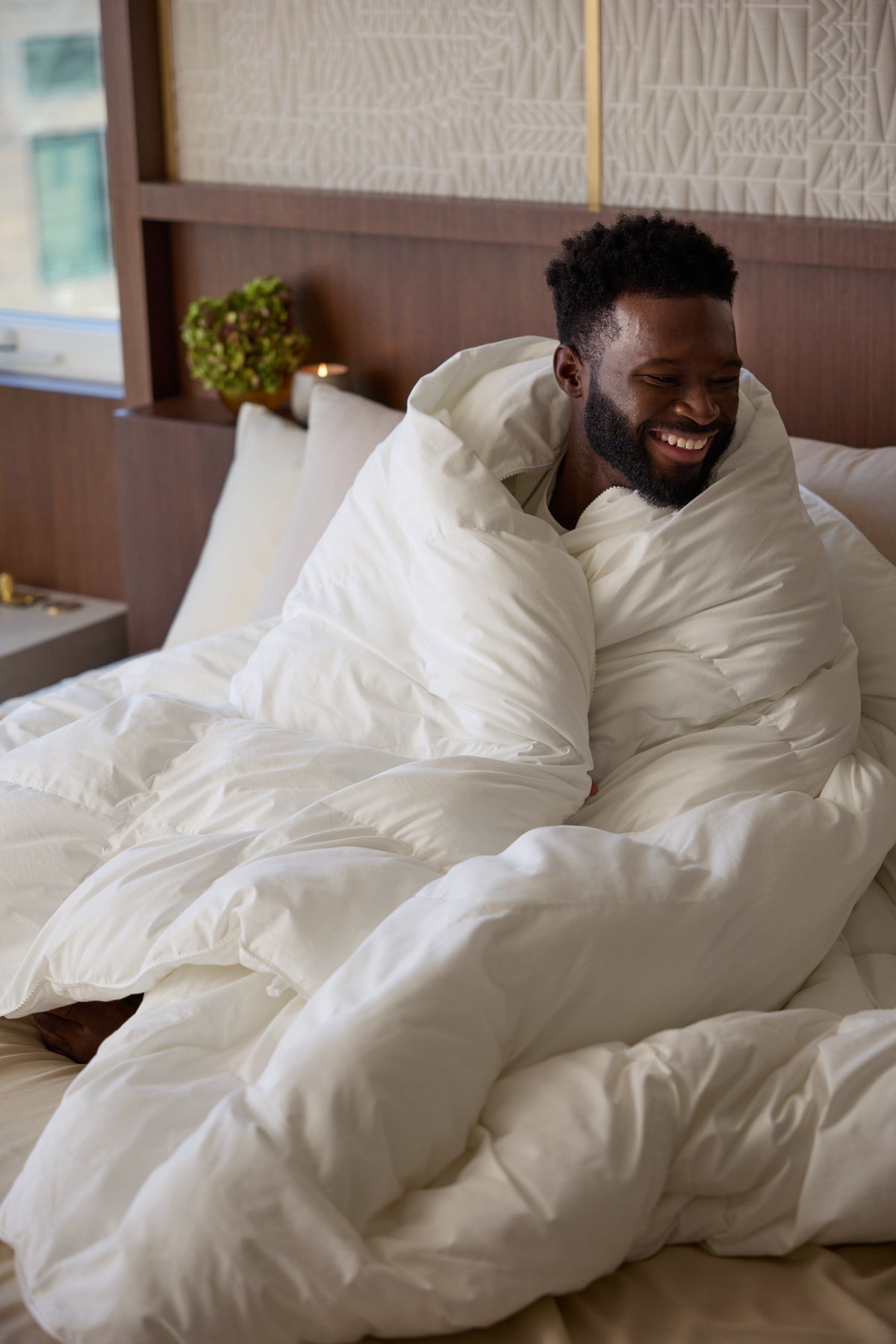 A man sits on a bed wrapped in the fluffy Cozy Earth Down Alternative Comforter, smiling and looking to the side in a cozy, well-lit bedroom. 